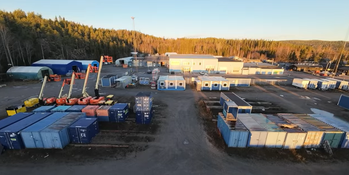 Drönarvy av ett kundcenter. Flera blå containrar och bodar står utspridda på en grusplan framför större anläggning.